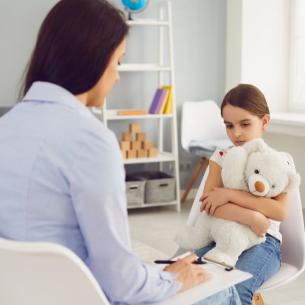 A young girl clutches a teddy bear while speaking with an adult seated opposite her.