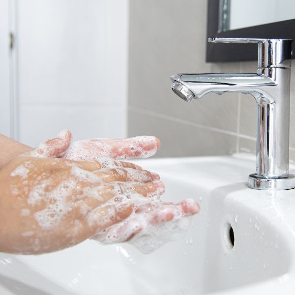 Hands covered in soap lather under a running faucet in a white bathroom sink, demonstrating proper handwashing.