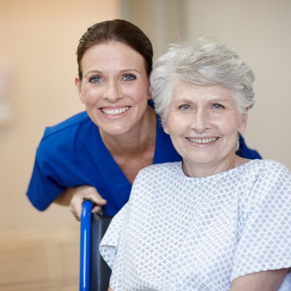 A smiling healthcare worker in blue scrubs stands behind a smiling senior female patient in a hospital gown sitting in a wheelchair.