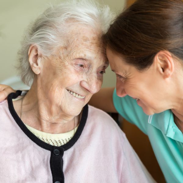 A smiling elderly woman with white hair and a pink cardigan gently touches foreheads with a younger woman in a teal top, both looking at each other with warmth and affection.
