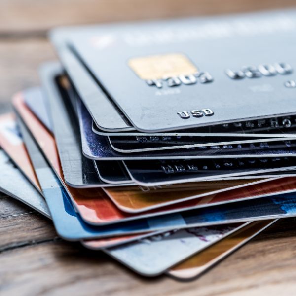 A close-up, low-angle shot of a stack of multiple credit and debit cards of various colors, with the top card partially showing its numbers and USD currency symbol. They are resting on a wooden surface.