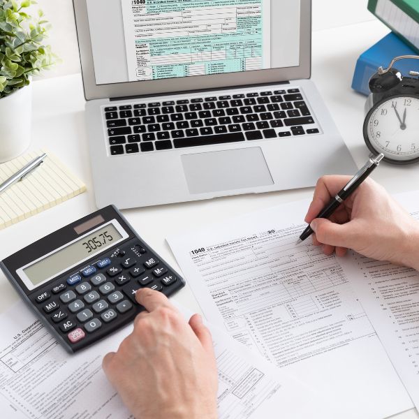A person’s hands working on a tax form with a laptop, calculator, and alarm clock nearby.