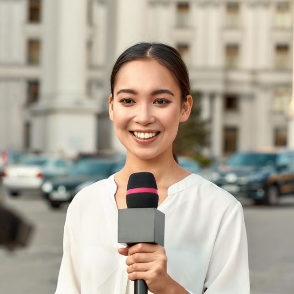 A smiling female journalist in a white shirt holds a microphone with a black and pink foam cover on a city street.